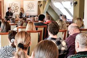 Pokaż zdjęcie: Grupa osób siedzi na drewnianych krzesłach w tej samej sali, uważnie słuchając lub obserwując przebieg wydarzenia. Wśród nich są zarówno mężczyźni, jak i kobiety, niektórzy z widoczną niepełnosprawnością lub trudnościami. Panuje skupiona, spokojna atmosfera. W tle widać ozdobne rzeźby i jasne światło wpadające przez okna. Na zdjęciu widnieje znak wodny z nazwiskiem fotografa – Paweł Lemiesiewicz.