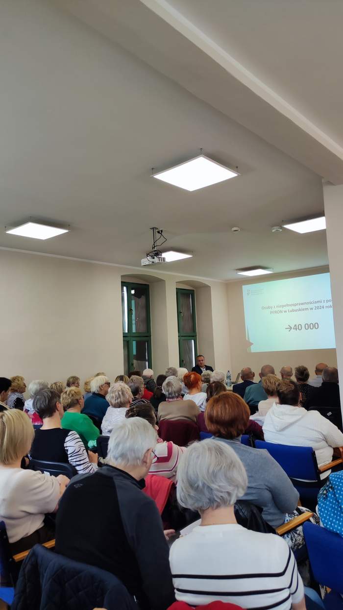 Pokaż zdjęcie: Na pierwszym planie uczestnicy spotkania, a w tle pracownik PFRON przedstawia prezentację