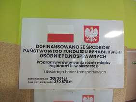 Pokaż zdjęcie: Tablica na górze flaga Polski i godło pod spodem z napis informujący o dofinansowaniu z PFRON.