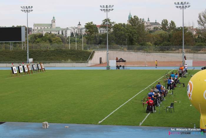 Pokaż zdjęcie: Zdjęcie stadionu, trwają zawody łucznicze, osoby siedzące na wózku inwalidzkim celują do tarczy
