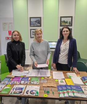 Pokaż zdjęcie: Na zdjęciu trzy kobiety – pracownice Oddziału Podlaskiego PFRON i Powiatowego Urzędu Pracy w Białymstoku – stojące za stołem pełnym materiałów informacyjnych i broszur. W tle widoczna jest jasna ściana z fotografiami oraz elementy wystroju wnętrza. Na stole znajdują się ulotki, długopisy i inne gadżety promocyjne związane z działalnością obu instytucji.