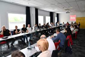 Pokaż zdjęcie: Sala konferencyjna. Przy ustawionych w dwóch rzędach stolikach siedzą uczestnicy spotkania.