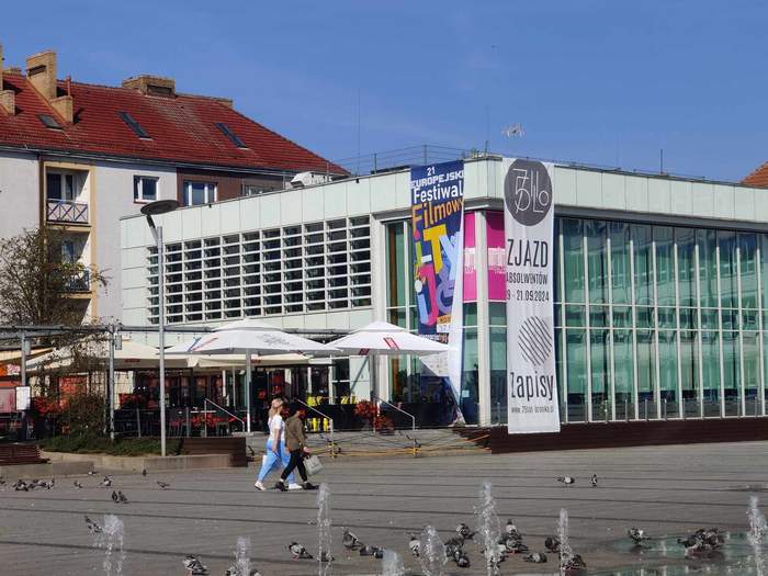 Pokaż zdjęcie: Biały budynek biblioteki w Koszalinie, gdzie odbywa się festiwal z flagami z logiem festiwalu 