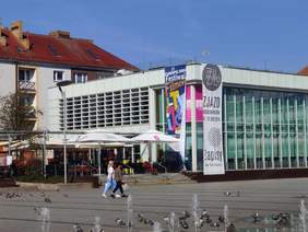 Pokaż zdjęcie: Biały budynek biblioteki w Koszalinie, gdzie odbywa się festiwal z flagami z logiem festiwalu 