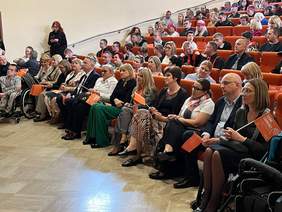 Pokaż zdjęcie: W sali widowiskowej Caritas siedzą zaproszeni goście, patrzą w kierunku sceny. W rękach trzymają pomarańczowe chorągiewki.