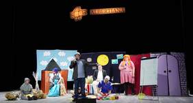 Pokaż zdjęcie: Scena Teatru z kolorową scenografią. Na scenie 7 aktorów z niepełnosprawnościami. Aktor na środku śpiewa do mikrofonu.  