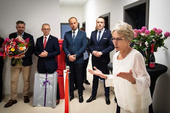 Pokaż zdjęcie: Kobieta w białej sukni w trakcie przemówienia i widać, że gestykuluje, a w tle inne osoby uczestniące w uroczystości 