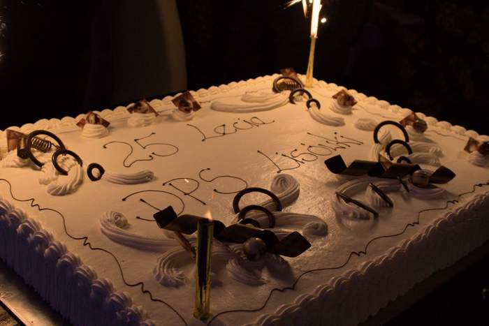 Pokaż zdjęcie: Tort w beżowym kolorze z napisem: „25 lecie DPS Lisówki”. Na obrzeżach tortu widoczne są dekoracje przestrzenne. Ustawione są na nim także świeczki zimne ognie, które zostały już zapalone.