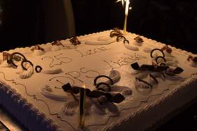 Pokaż zdjęcie: Tort w beżowym kolorze z napisem: „25 lecie DPS Lisówki”. Na obrzeżach tortu widoczne są dekoracje przestrzenne. Ustawione są na nim także świeczki zimne ognie, które zostały już zapalone.