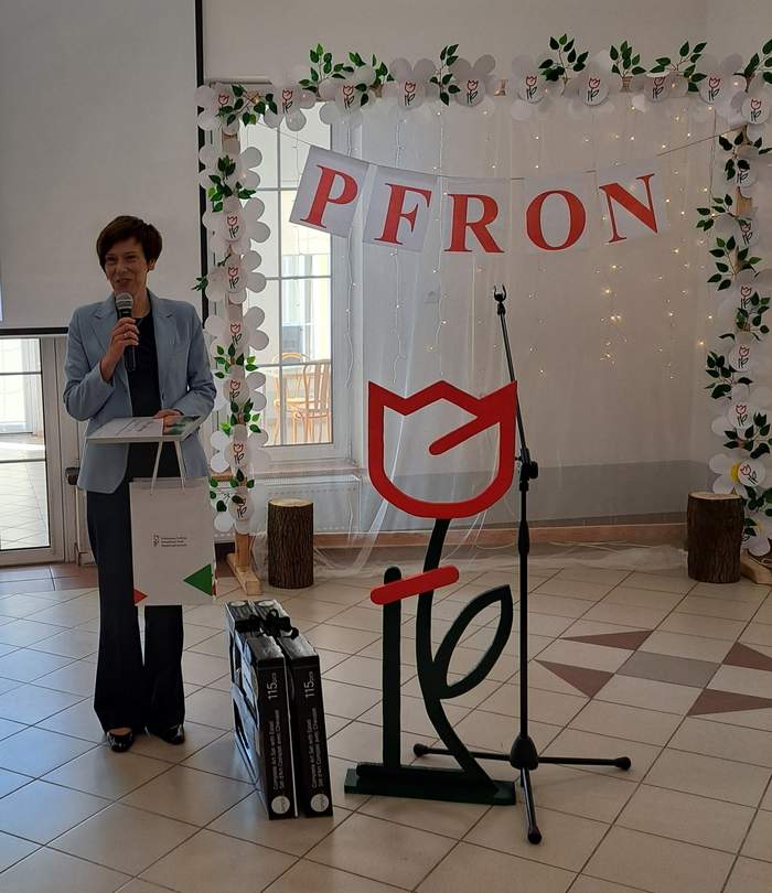 Pokaż zdjęcie: Kobieta z mikrofonem trzyma w ręce dyplom i torbę z logo PFRON. Obok kobiety, na podłodze, znajdują się dwa zestawy artystyczne oraz duży tulipan z logotypu PFRON wykonany ze styropianu. W tle drewniana brama udekorowana papierowymi tulipanami.