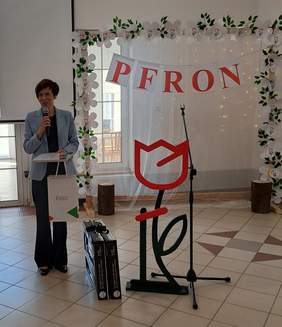 Pokaż zdjęcie: Kobieta z mikrofonem trzyma w ręce dyplom i torbę z logo PFRON. Obok kobiety, na podłodze, znajdują się dwa zestawy artystyczne oraz duży tulipan z logotypu PFRON wykonany ze styropianu. W tle drewniana brama udekorowana papierowymi tulipanami.