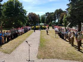 Na terenie zielonym stoją harcerze. Ustawieni są w dwóch długich rzędach. Na środku stoi mikrofon. W tle widać budynki i drzewa. 
