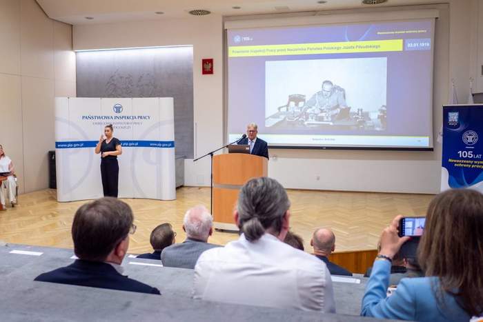 Pokaż zdjęcie: Na sali konferencyjnej siedzą uczestnicy i słuchają wystąpienia Andrzeja Bugalskiego. W tle wyświetlona jest prezentacja ze zdjęciem Józefa Piłsudskiego. Na sali na tle bannera Państwowej Inspekcji Pracy kobieta tłumaczy wystąpienie na język m