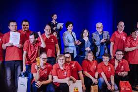 Pokaż zdjęcie: Na scenie znajduje się grupa teatralna, która zwyciężyła w festiwalu Albertiana. Aktorzy trzymają w dłoniach nagrody, torby upominkowe, statuetkę i dyplom okolicznościowy. Wszyscy są ubrani w czerwone koszulki. 