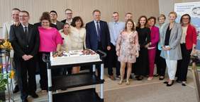 Pokaż zdjęcie: Grupa osób pozuje do wspólnego zdjęcia podczas obchodów 30-lecia Warsztatu Terapii Zajęciowej TPD w Suwałkach. Na pierwszym planie stoi duży tort udekorowany owocami i świeczkami. W tle widać uczestników warsztatu, organizatorów oraz zaproszonych gości.