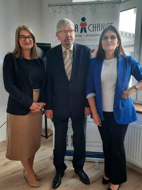 Pokaż zdjęcie: Na tle baneru Fundacji Szansa stoją trzy osoby: dwie kobiety i mężczyzna. Panie są elegancko ubrane, a pan jest w garniturze. Wszyscy się uśmiechają. 