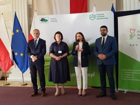 4 osoby, dwie kobiety pośrodku, dwóch mężczyzn po bokach, ubrani oficjalnie, stoją na tle banerów i flag, jedna z kobiet przemawia.