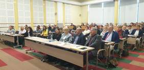 Pokaż zdjęcie: Na zdjęciu widać salę konferencyjną z kilkudziesięcioma osobami siedzącymi przy stołach. Na stołach znajdują się dokumenty, butelki z wodą i plastikowe kubki. Atmosfera sugeruje na formalne spotkanie, takie jak szkolenie lub wykład.