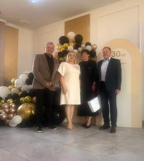 Pokaż zdjęcie: Cztery osoby stoją na tle dekoracji z balonów w jasnym pomieszczeniu z podłogą z szarych płytek. Od lewej stoją: mężczyzna w beżowej marynarce i ciemnej koszuli, kobieta z jasnymi włosami do ramion w prostej, jasnej sukience, kobieta z krótkimi, ciemnymi włosami w ciemnej sukience, trzymająca jasną torebkę prezentową, oraz mężczyzna w ciemnej marynarce i jasnej koszuli. Za nimi znajduje się gęsta kompozycja z balonów w kolorach złotym, białym, czarnym i perłowym. Po prawej stronie tła widoczny jest zaokrąglony panel z napisem trzydzieści lat.