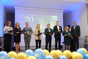 Pokaż zdjęcie: Grupa ośmiu elegancko ubranych osób stoi w rzędzie na scenie, trzymając w dłoniach czerwoną róże oraz granatową teczkę. Za nimi, na dużym ekranie w niebieskim świetle, wyświetla się napis „20-lecie” z okazji jubileuszu. Przed sceną, na całej jej szerokości, ułożone są gęsto żółte i niebieskie balony.