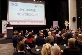 Pokaż zdjęcie: Sala konferencyjna wypełniona uczestnikami siedzącymi przy okrągłych stolikach, na scenie mównica z przemawiającym mężczyzną i panel gości. Na ekranie napis „Spotkanie Noworoczne”, logotypy Gminy Szemud i organizacji partnerskiej.