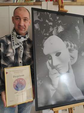 Osoba stojąca obok dużej, czarno-białej fotografii umieszczonej na sztaludze, trzymająca w dłoni dyplom za zdobycie pierwszego miejsca w konkursie plastycznym.