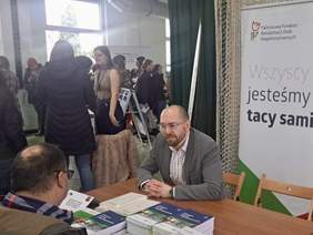 Pokaż zdjęcie: Mężczyzna w marynarce rozmawia z odwiedzającym przy stoliku PFRON, na którym leżą broszury i dokumenty. W tle tłum ludzi, baner z hasłem „Wszyscy jesteśmy tacy sami” oraz logo Państwowego Funduszu Rehabilitacji Osób Niepełnosprawnych.