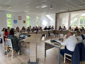 Pokaż zdjęcie: Jasna sala konferencyjna, z dużymi oknami. Stoły  konferencyjne w kolorze jasnego drewna połączone w duży prostokąt, za którym siedzi kilkudziesięciu ludzi. Między nimi stoi mównica.  