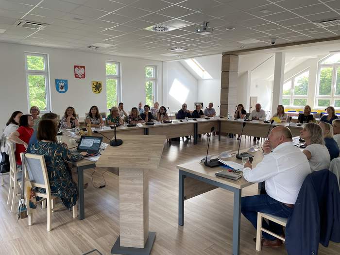 Pokaż zdjęcie: Jasna sala konferencyjna, z dużymi oknami. Stoły  konferencyjne w kolorze jasnego drewna połączone w duży prostokąt, za którym siedzi kilkudziesięciu ludzi. Między nimi stoi mównica.  