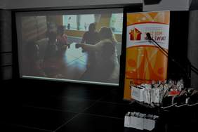 Pokaż zdjęcie: Po lewej stronie znajduje się duży ekran, na którym widnieje kadr z filmu. Z kolei po prawej stronie ustawiony jest baner Stowarzyszenia Na Rzecz Osób Niepełnosprawnych „Nasz Dom - Nasz Świat” z Leszna oraz nagrody rzeczowe i statuetki.