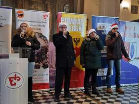 Pokaż zdjęcie: W kościele znajdują się cztery osoby: dwie kobiety i dwóch mężczyzn. Wszyscy trzymają w dłoniach mikrofony, do których śpiewają. Na głowach mają czapki świętego Mikołaja. 