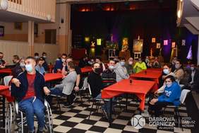 Pokaż zdjęcie: Na dużej sali widowiskowej siedzą osoby przy stolikach z czerwonymi obrusami. W tle na scenie są ustawione obrazy i rękodzieło wykonane przez uczestników placówki.