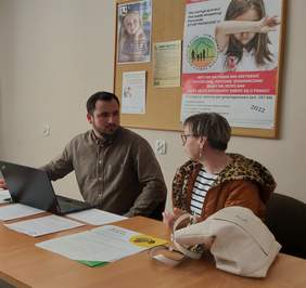 Pokaż zdjęcie: Mężczyzna i kobieta siedzą przy stole. Przed nimi laptop oraz ulotki. Znajdują się w pomieszczeniu z jasnymi ścianami. Za nimi tablica korkowa z umieszczonymi na niej plakatami dotyczącymi działalności Powiatowego Centrum Pomocy Rodzinie w Piszu.