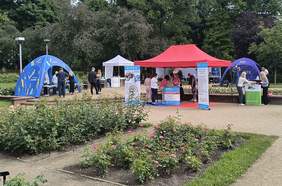 Pokaż zdjęcie: Stoiska informacyjne ustawione w ogrodzie różanym. Kolorowe namioty (niebieski, czerwony i biały) należą do różnych instytucji i organizacji, m.in. ZKKZ, Pomorze Zachodnie i NFZ. Przed stoiskami stoją ludzie rozmawiający z przedstawicielami, na pierwszym planie rabaty różane.