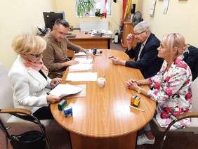 Pokaż zdjęcie: Przy owalnym stole siedzą dwie kobiety i dwóch mężczyzn. Na stole leżą dokumenty, pieczątki, długopisy, telefony. 