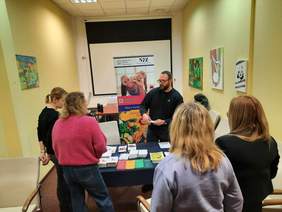 Pokaż zdjęcie: W pokoju znajduje sie pięć osób: cztery kobiety i jeden mężczyzna. Wszyscy stoją przy stoisku profilaktyki i promocji zdrowia. Pan tłumaczy coś paniom i podaje im materiały reklamowe. 