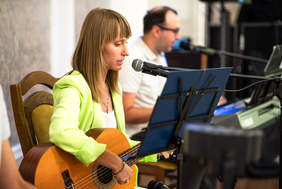 Mężczyzna i kobieta siedzą za mikrofonami, śpiewają, kobieta jest na pierwszym planie, gra na gitarze