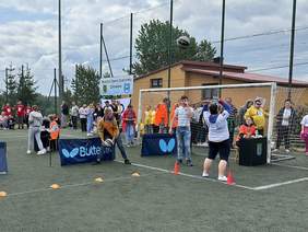 Pokaż zdjęcie: Kobieta rzuca piłką do tyłu. Przed nią bramka piłkarska za którą stoją inni zawodnicy. Za nią wyznaczone punkty plastikowymi pachołkami. Na siace ochronnej napis Warsztat Terapii Zajęciowej Chmielno