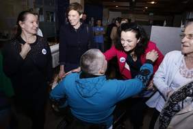 Pokaż zdjęcie: W wąskim pomieszczeniu, na pierwszym planie znajduje się pięć kobiet. Jedna z nich wita się z mężczyzną poruszającym się na wózku, mając dłoń położoną na jego ramieniu. W sali możemy dostrzec także tort ustawiony na stoliku oraz grupę pięciu innych osób, które znajdują się z tyłu sali.