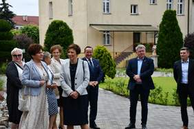 Pokaż zdjęcie: Grupa ośmiu osób stoi na chodniku. W tle widoczne są dwa budynki, trawnik oraz drzewa i krzewy, w przewadze iglaste.