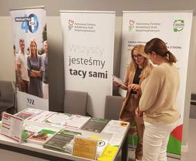 Pokaż zdjęcie: Kobieta stoi za stolikiem na tle baneru PFRON oraz CIDON i udziela informacji kobiecie, która przyszła do ZUS na Dzień Osób z Niepełnosprawnością. Na stoliku ułożone są materiały informacyjne promujące programy i zadania PFRON.