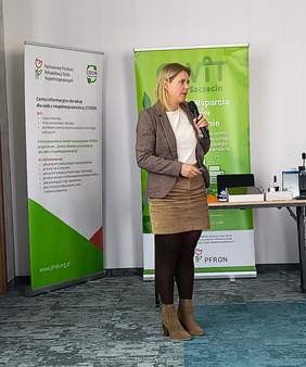 Pokaż zdjęcie: Na zdjęciu widzimy Annę Podgórą, Rehamanagera przedstawiającą prezentację na temat działań rehamanagera. W tle rollup PFRON i CODN w kolorach biało zielonych i zielony rollup OWiT. Widoczne również sprzęty wspomagające dla ON na stoliku. 