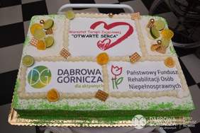 Pokaż zdjęcie: Tort w kształcie prostokąta ozdobiony plastrami cytryny i ciastek. Na górze napis warsztat terapii zajęciowej Otwarte Serca, poniżej z lewej strony napis Dąbrowa Górnicza dla aktywnych, z prawej logo PFRON.