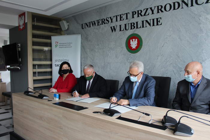 Pokaż zdjęcie: W sali konferencyjnej przy stolw siedż trzej mężczyźni oraz kobieta.Dwaj z nich podpisują dokumenty