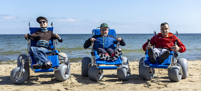 Pokaż zdjęcie: Trzy osoby siedzą na niebieskich wózkach plażowych z szerokimi kołami ustawionych na piaszczystej plaży. W tle widoczne jest morze i jasne niebo. Wszystkie osoby zwrócone są przodem do obiektywu.