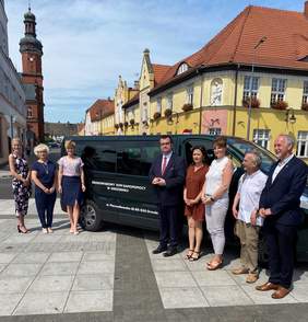 Pokaż zdjęcie: Dyrektor Oddziału Lubuskiego wraz z pozostałymi uczestnikami w trakcie uroczystości przekazania busa na tle nowego auta dla ŚDS w Drezdenku na rynku w Drezdenku