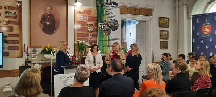 Pokaż zdjęcie: Zdjęcie czterech kobiet przy mównicy, jedna z nich mówi do mikrofonu, trzyma kwiaty, obok stoją trzy kobiety, jedna z nich również trzyma bukiet kwiatów w ręku.