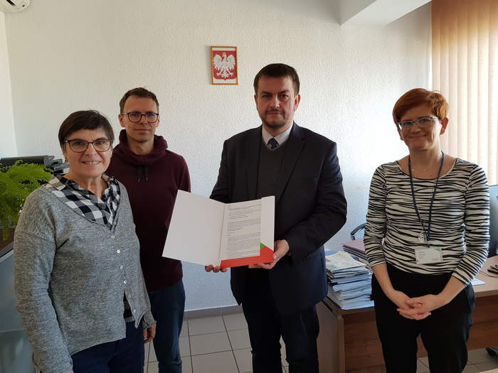 Pokaż zdjęcie: Dyrektor Oddziału wraz pracownikiem oraz przedstawicielami Stowarzyszenia stoją obok siebie z podpisaną umową w gabinecie Dyrektora, wszyscy są uśmiechnięci 