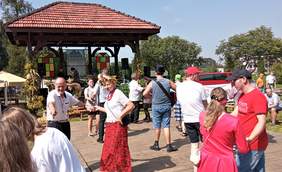 Pokaż zdjęcie: Zabawa dożynkowa w plenerze. Na pierwszym planie widoczna grupa tańczących osób w różnym wieku. W tle widoczna scena.