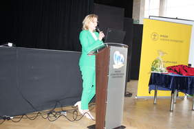 Pokaż zdjęcie: Przy mównicy stoi kobieta ubrana w zielony garnitur.